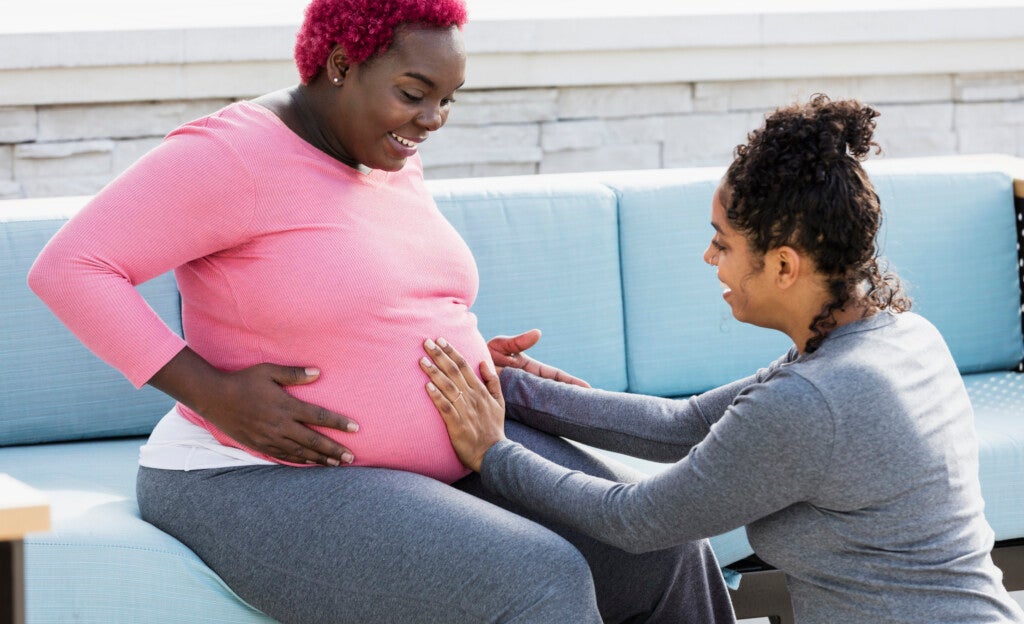 A pregnant African-American woman with her doula or birth support coach. The expectant mother, who had pink hair and is wearing a pink shirt, is sitting outdoors on a patio sofa smiling. The doula, a mixed race woman, is kneeling in front of her, touching her round abdomen. Both women are in their 30s.