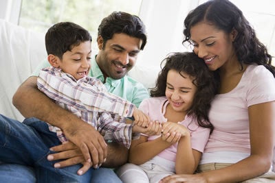 A family is gathered together on a couch; a father holds a young son who playfully gestures at his sister and mother.